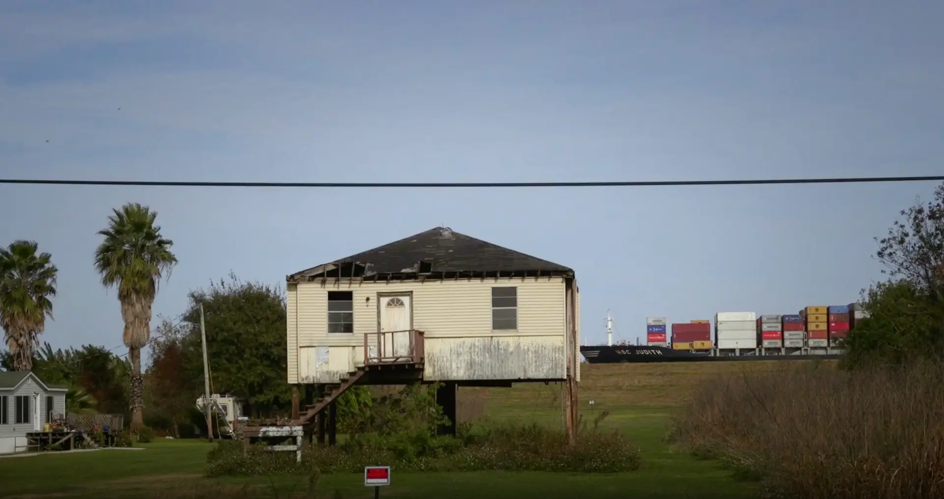 Raised house on stilts beside a levee as a container ship passes